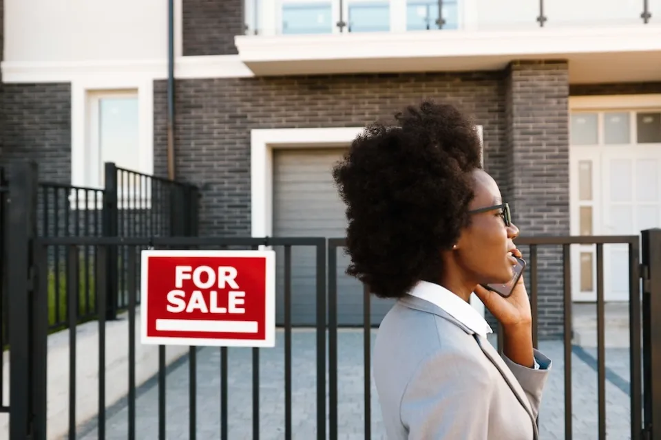 Woman realtor or home buyer client wearing blazer walking past urban condo with red and white For Sale sign while talking on phone.
